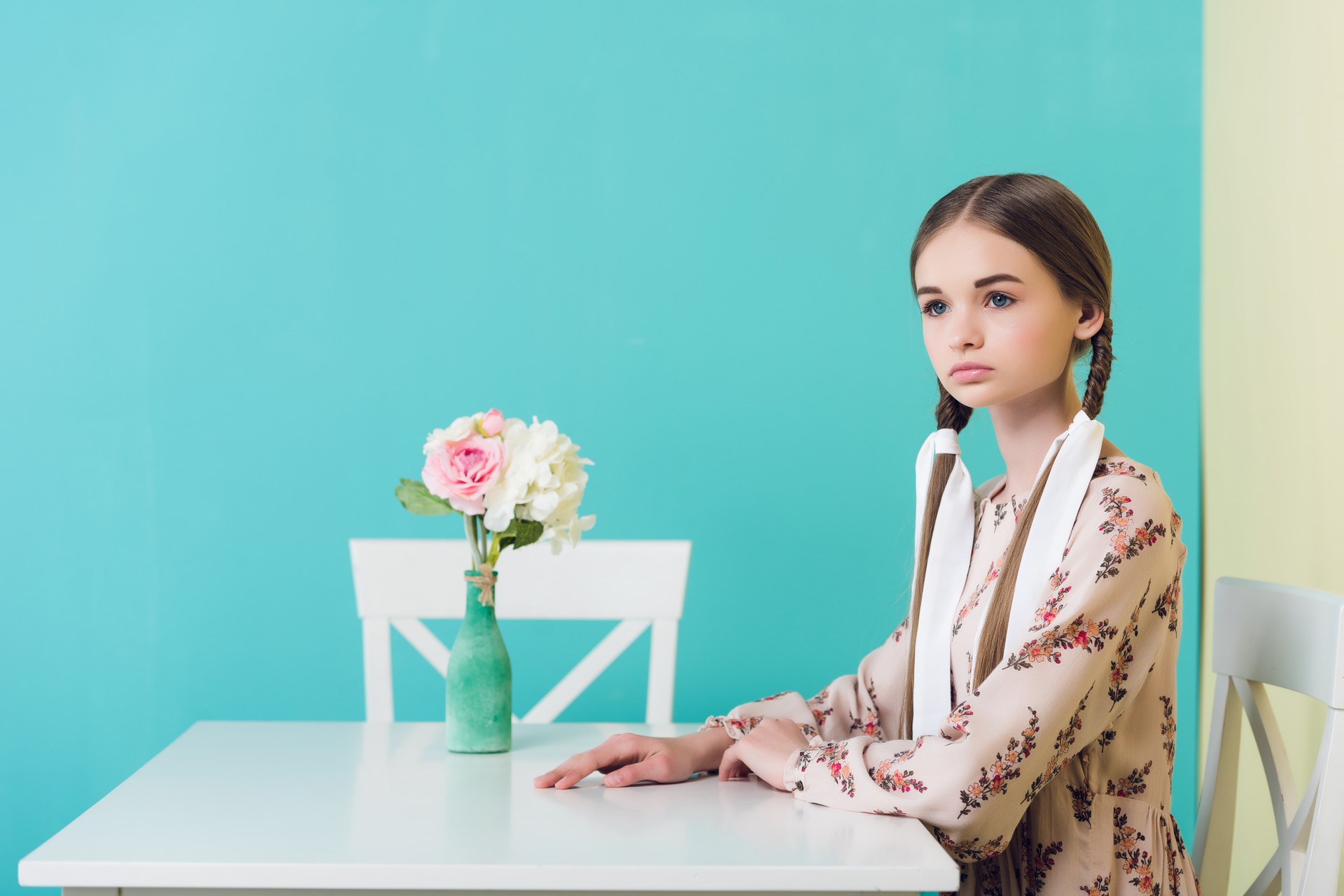 teen girl with braids in summer dress sitting at table with flowers, on blue